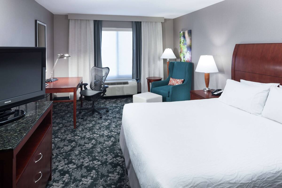 Une chambre d’hôtel avec un lit, un bureau avec une télévision, une chaise, une lampe et une fenêtre avec des rideaux.