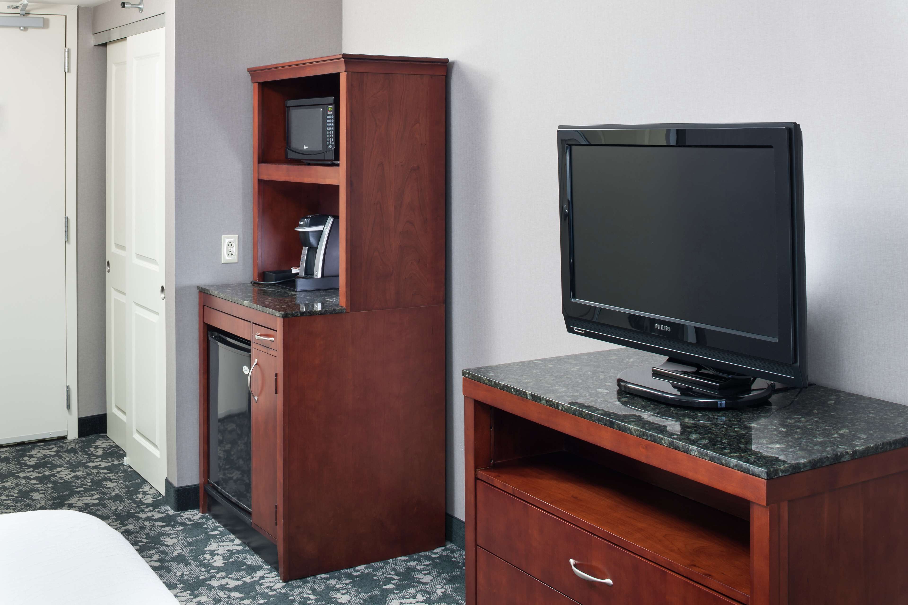 A compact hotel room with a wooden desk, a microwave, a television, and a bed with a white headboard.