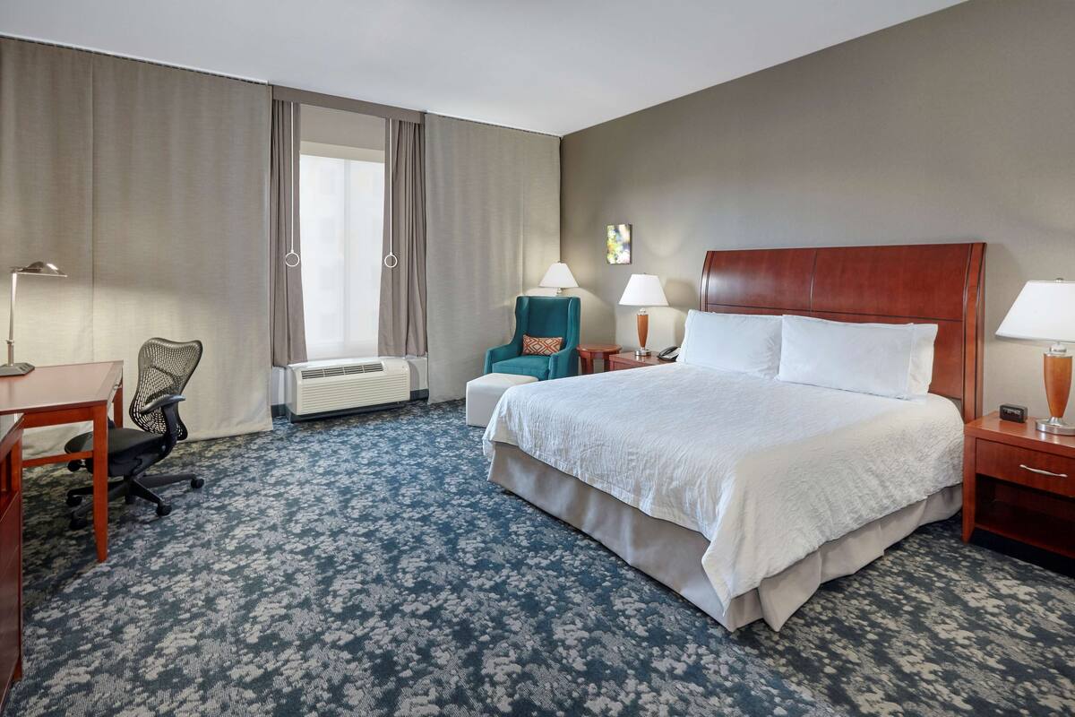 Une chambre d’hôtel avec un grand lit, un bureau, une chaise, deux lampes et une fenêtre avec des rideaux.