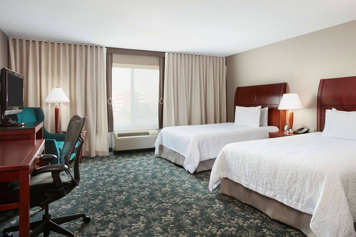 Une chambre d’hôtel avec deux lits, un bureau avec une chaise, une télévision et une fenêtre avec des rideaux.