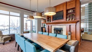 A spacious dining area with a long wooden table and blue upholstered chairs, a fireplace, and a television mounted above it. - Hilton Garden Inn Naperville/Warrenville (Warrenville)