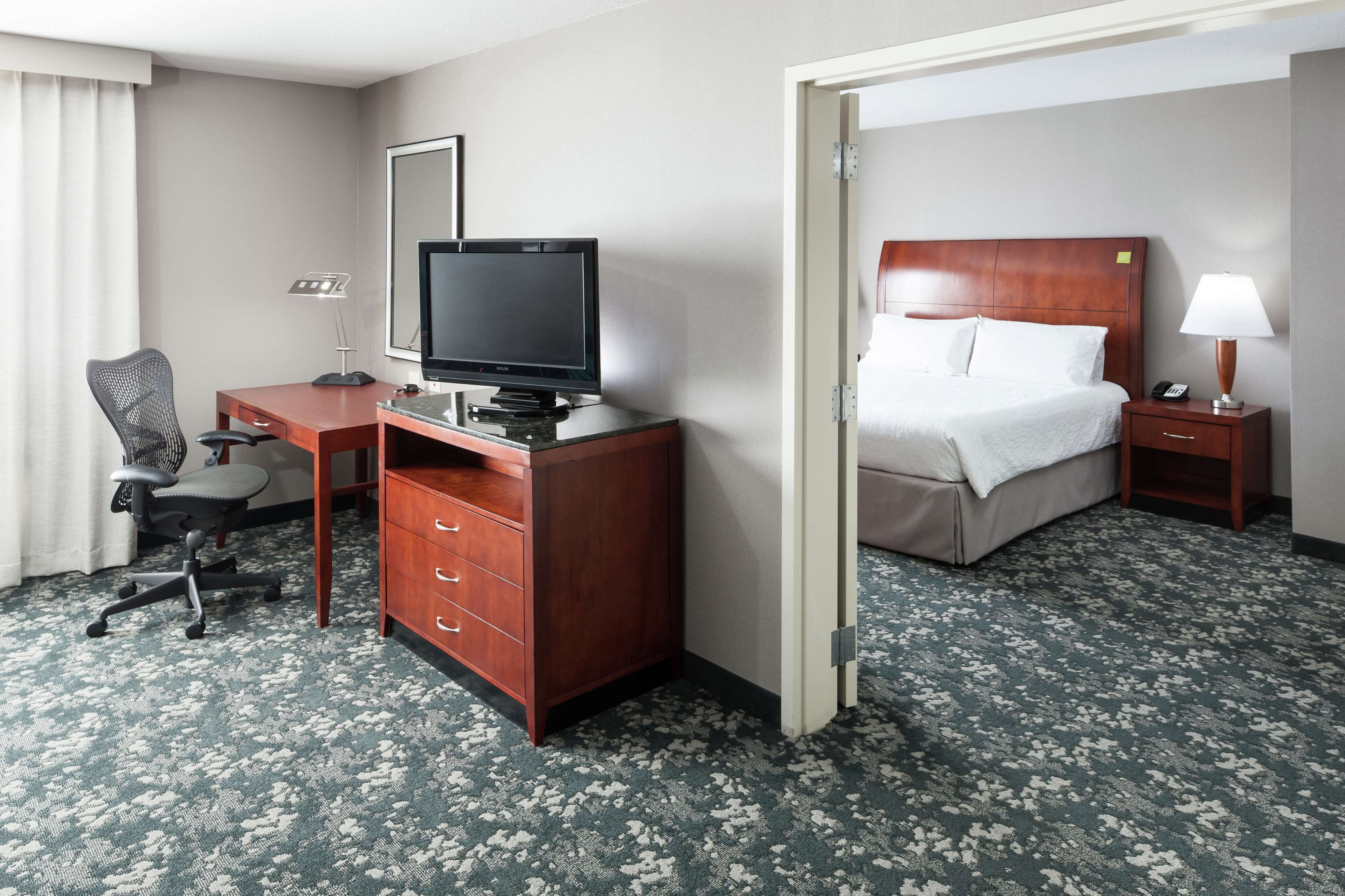 A hotel room with a bed, a desk with a television, a chair, a nightstand with a lamp, and a carpeted floor.
