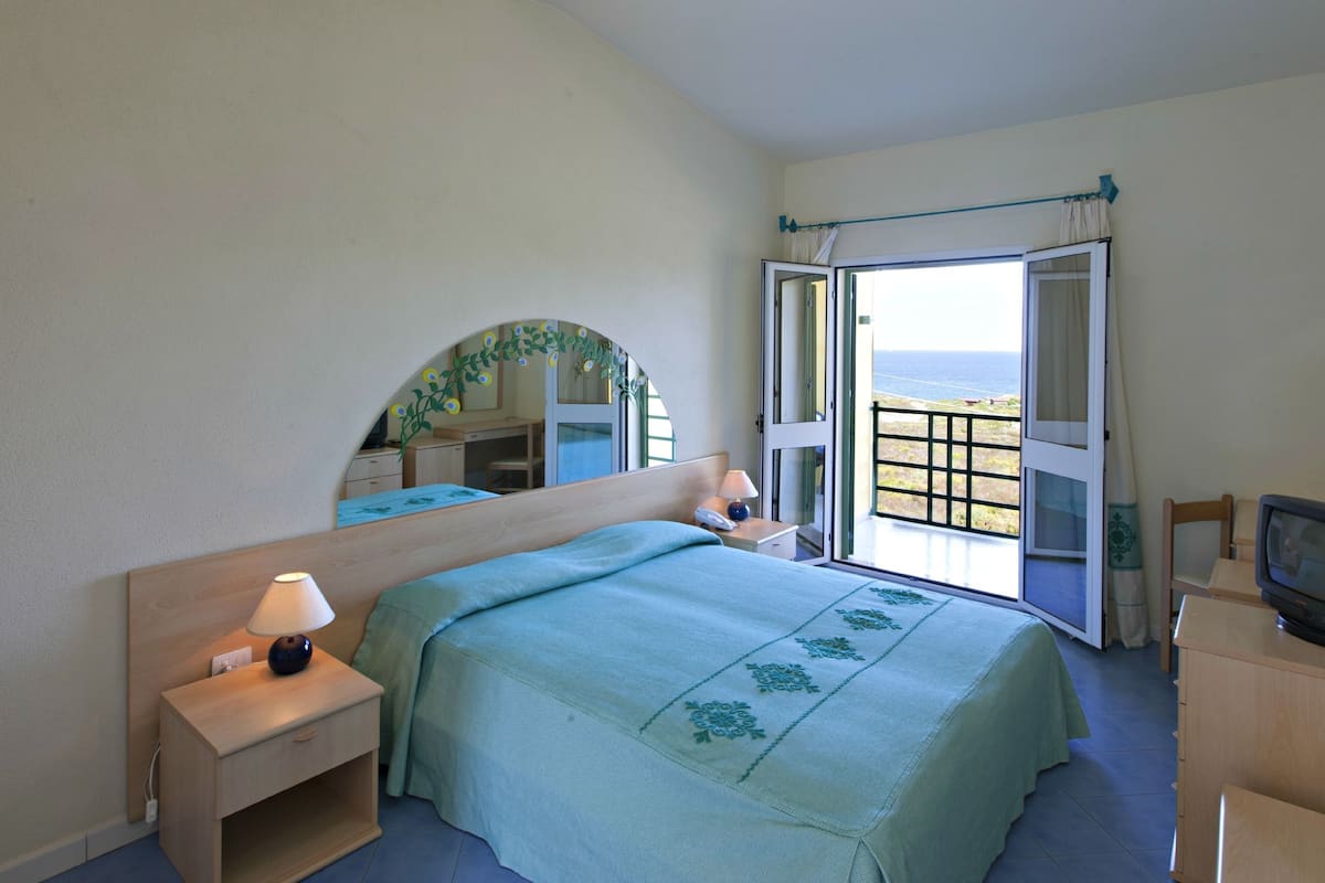 A bedroom with a bed, a nightstand, a TV, and an open door leading to a balcony with a view.