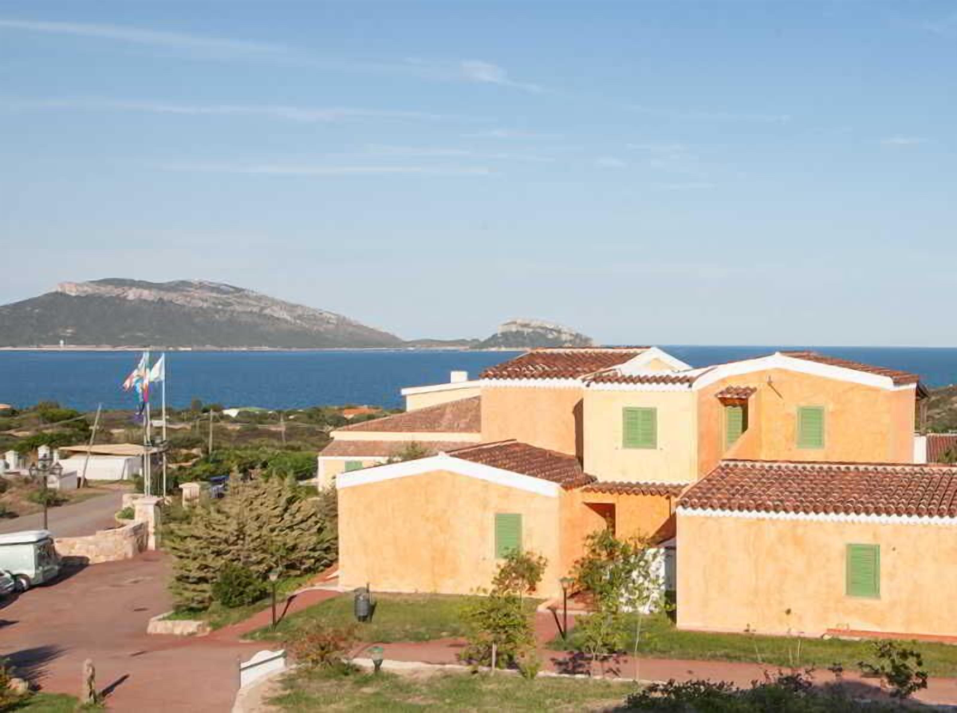 Un quartier résidentiel en bord de mer, avec des bâtiments aux toits de tuiles rouges, une vue dégagée sur la mer et une chaîne de montagnes à l’horizon.