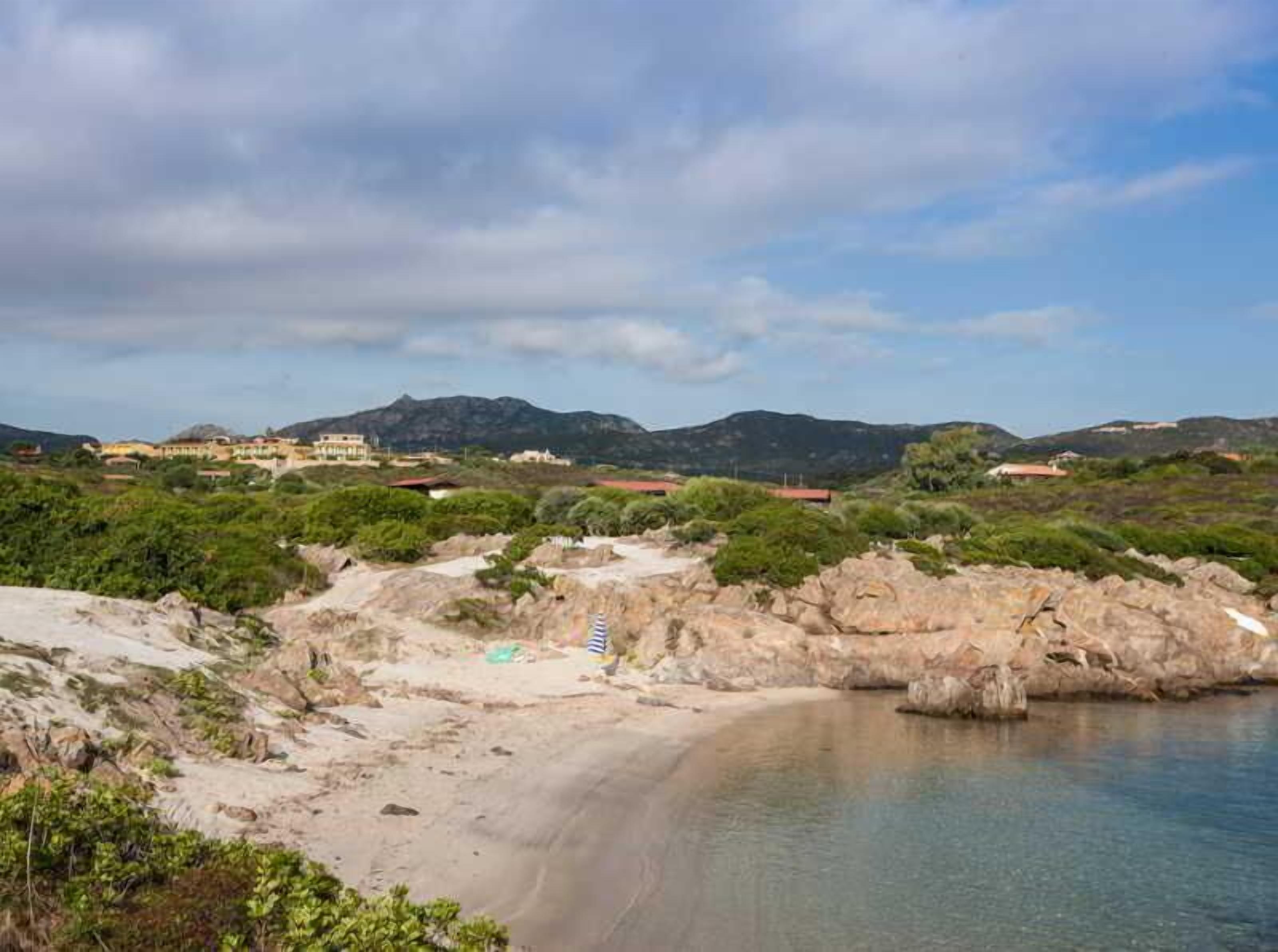 A coastal landscape with a sandy beach, rocky outcrops, and a mountain range in the background.