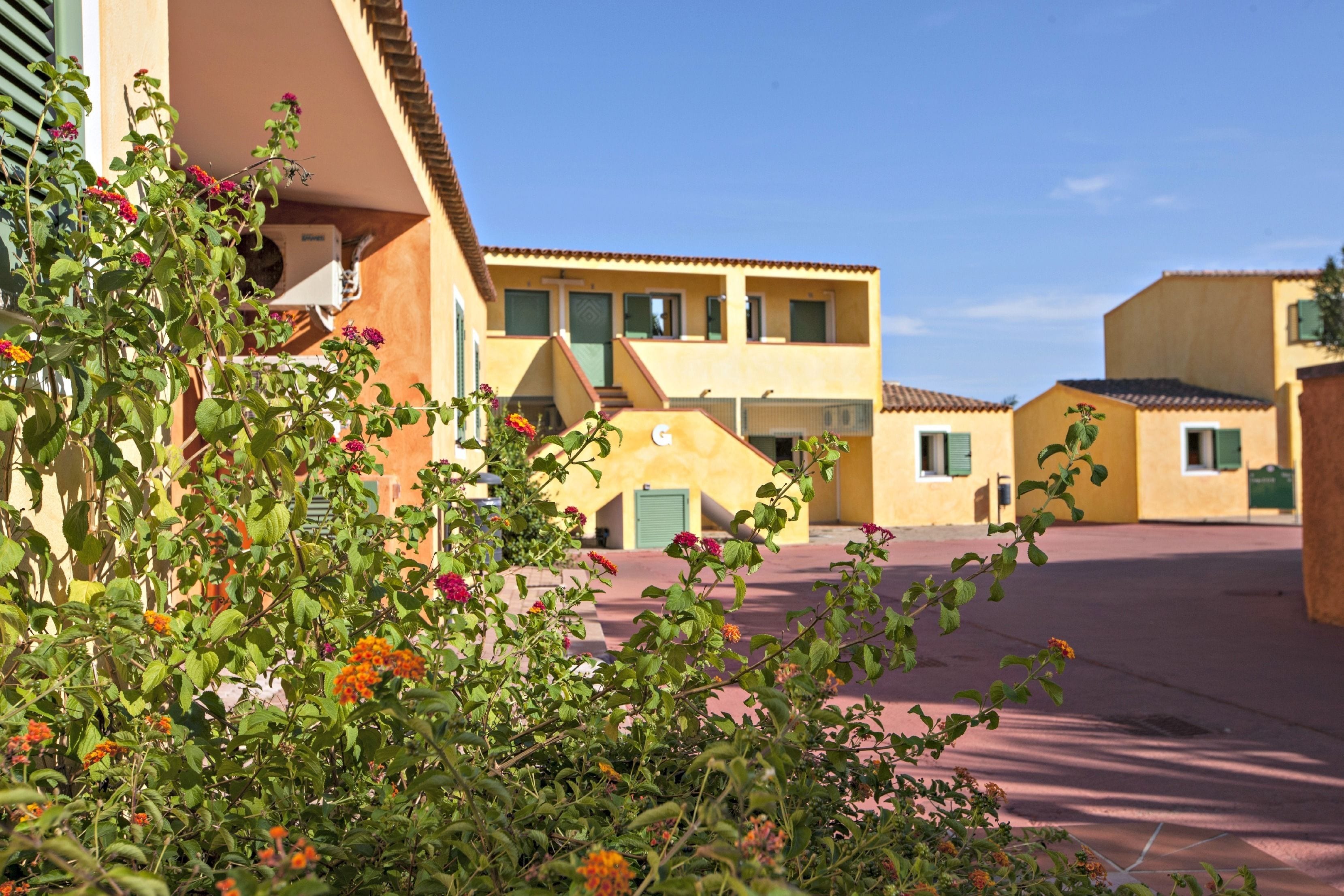 A courtyard with buildings featuring a yellow facade and green shutters, surrounded by lush greenery and flowering plants.