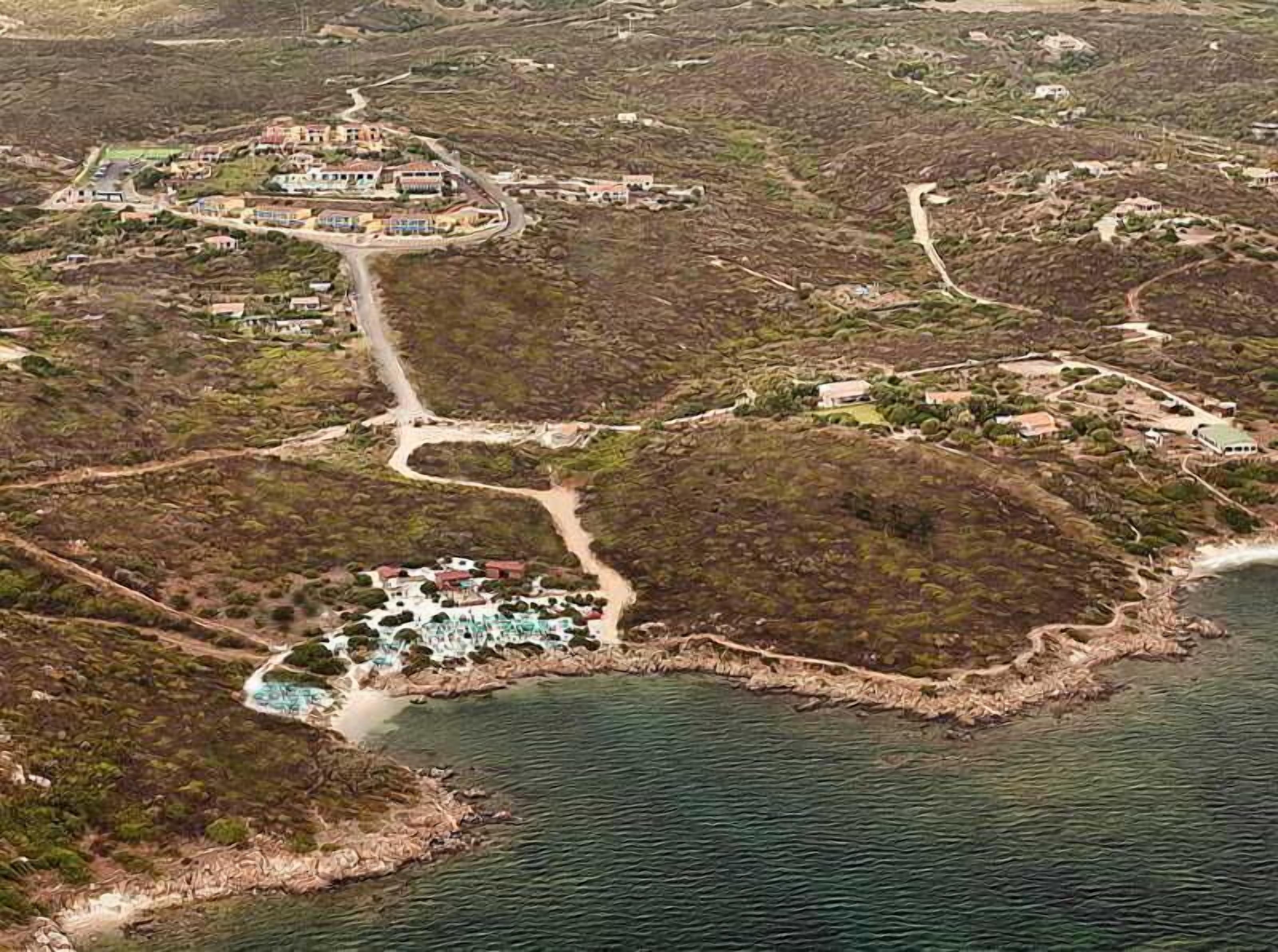 Vista aérea de un complejo turístico costero con varias construcciones, un área central para nadar y un paisaje natural circundante.