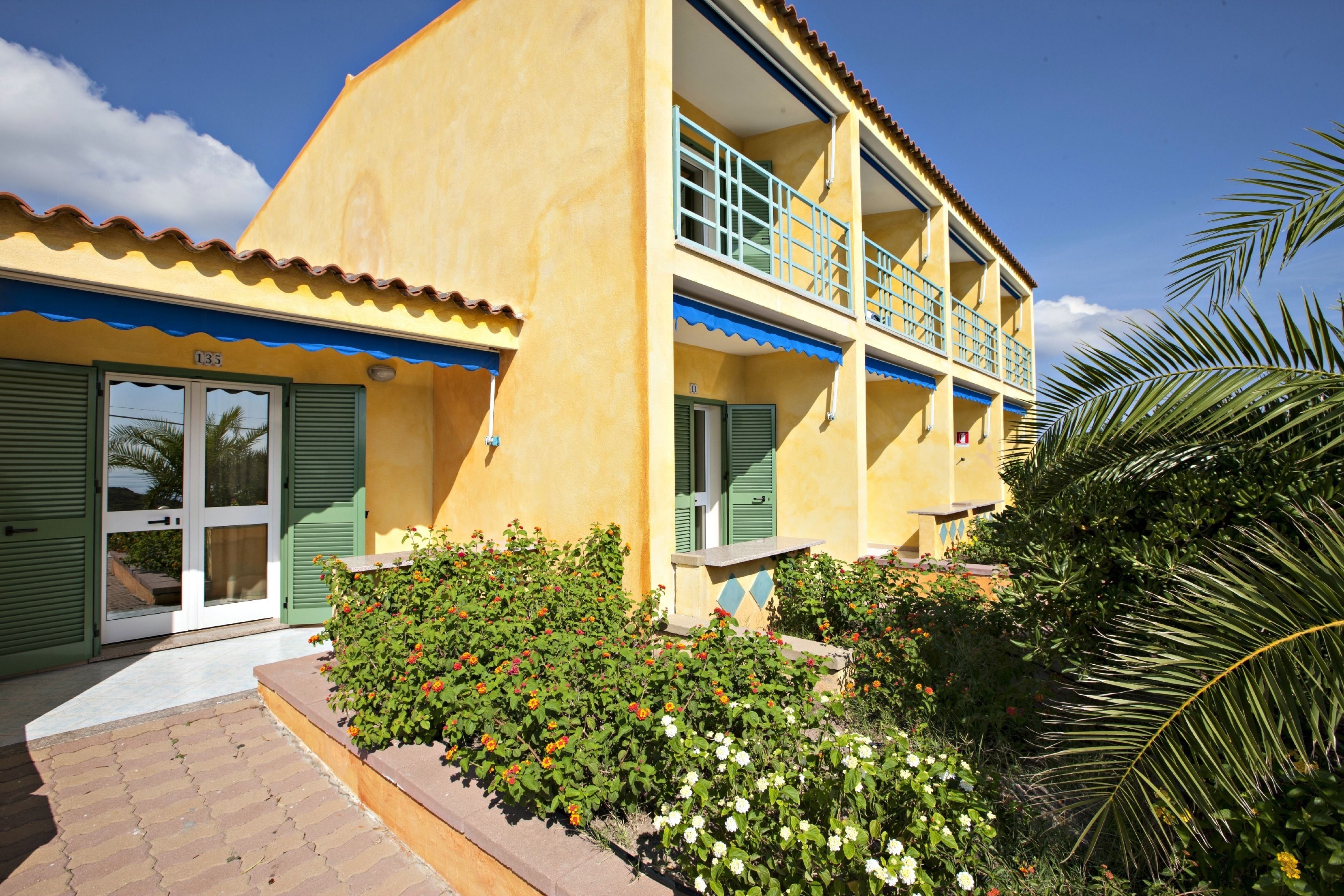 A two-story building with a yellow facade, green shutters, and a balcony with blue railings.