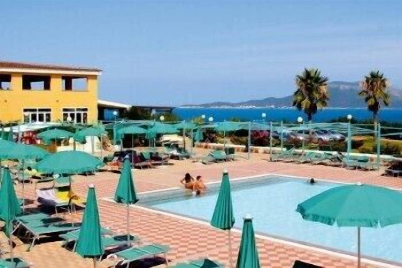 A pool area with lounge chairs and umbrellas, a building with multiple windows, and a view of the sea.