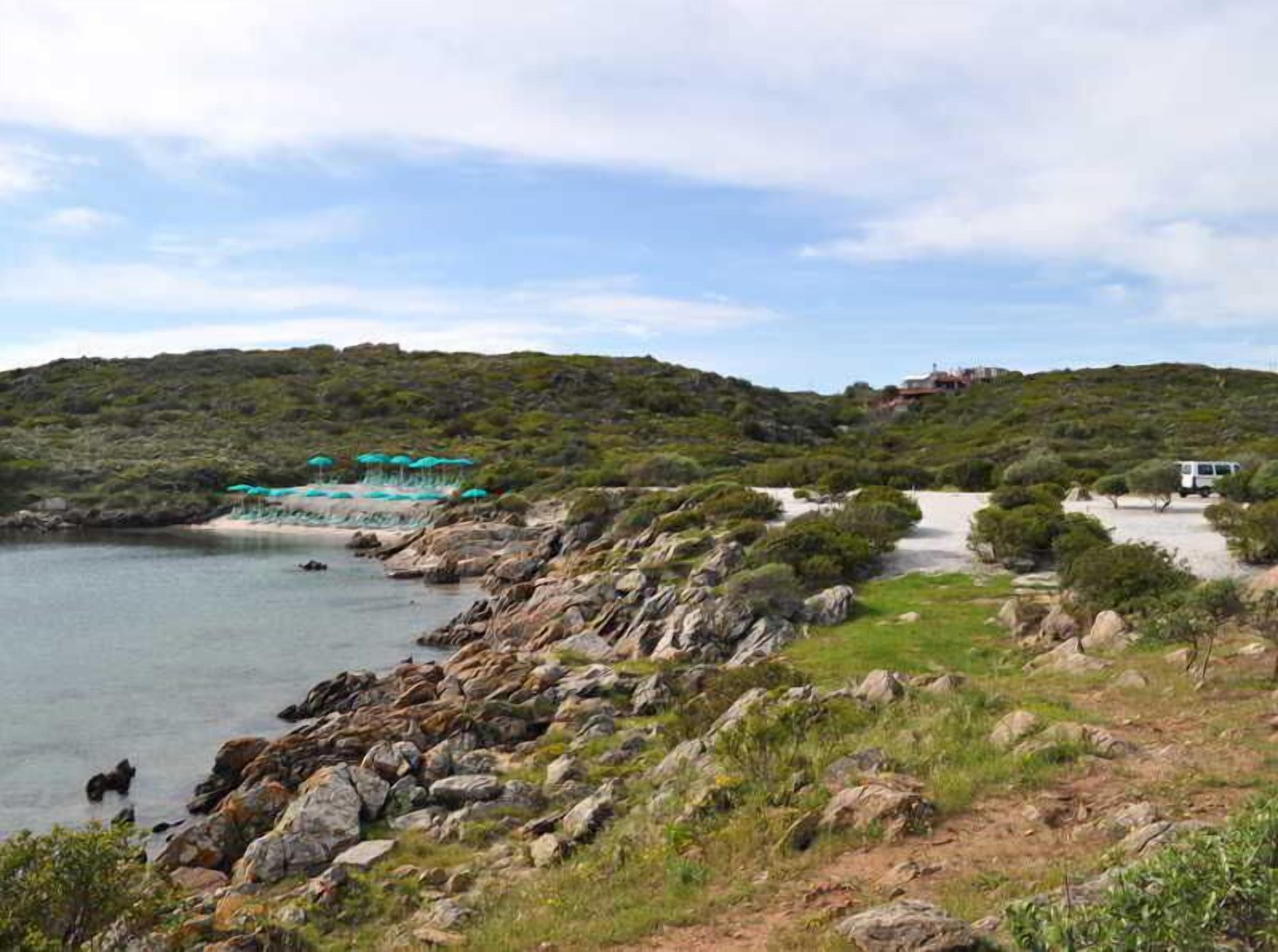 A coastal landscape with a rocky shoreline, a small beach area with umbrellas, and a path leading to a building on a hill.