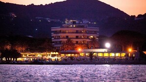Un hotel de varios pisos con zona de piscina, palmeras y montañas al fondo.
