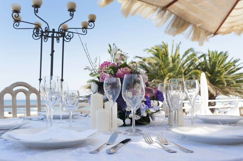 A table set for a formal outdoor dining experience with a decorative centerpiece, candles, and a coastal view.
