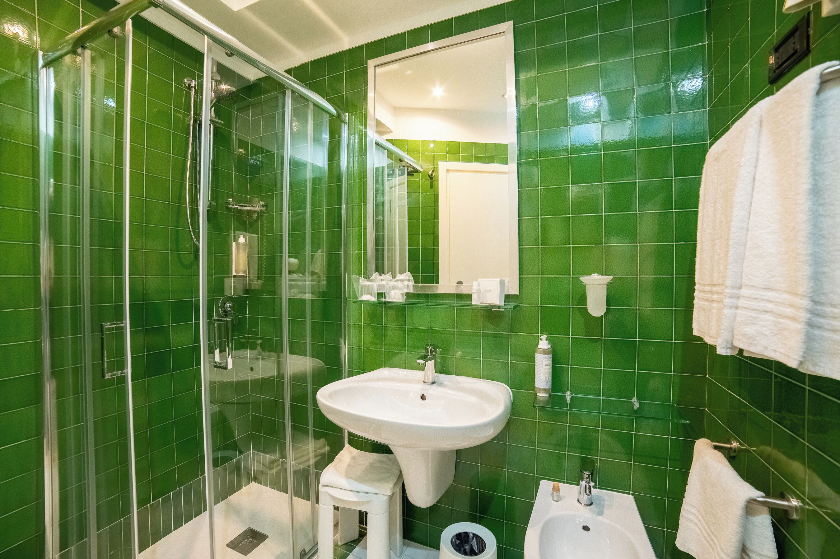 Un baño con azulejos verdes, cabina de ducha de vidrio, lavabo, inodoro y un perchero para toallas.