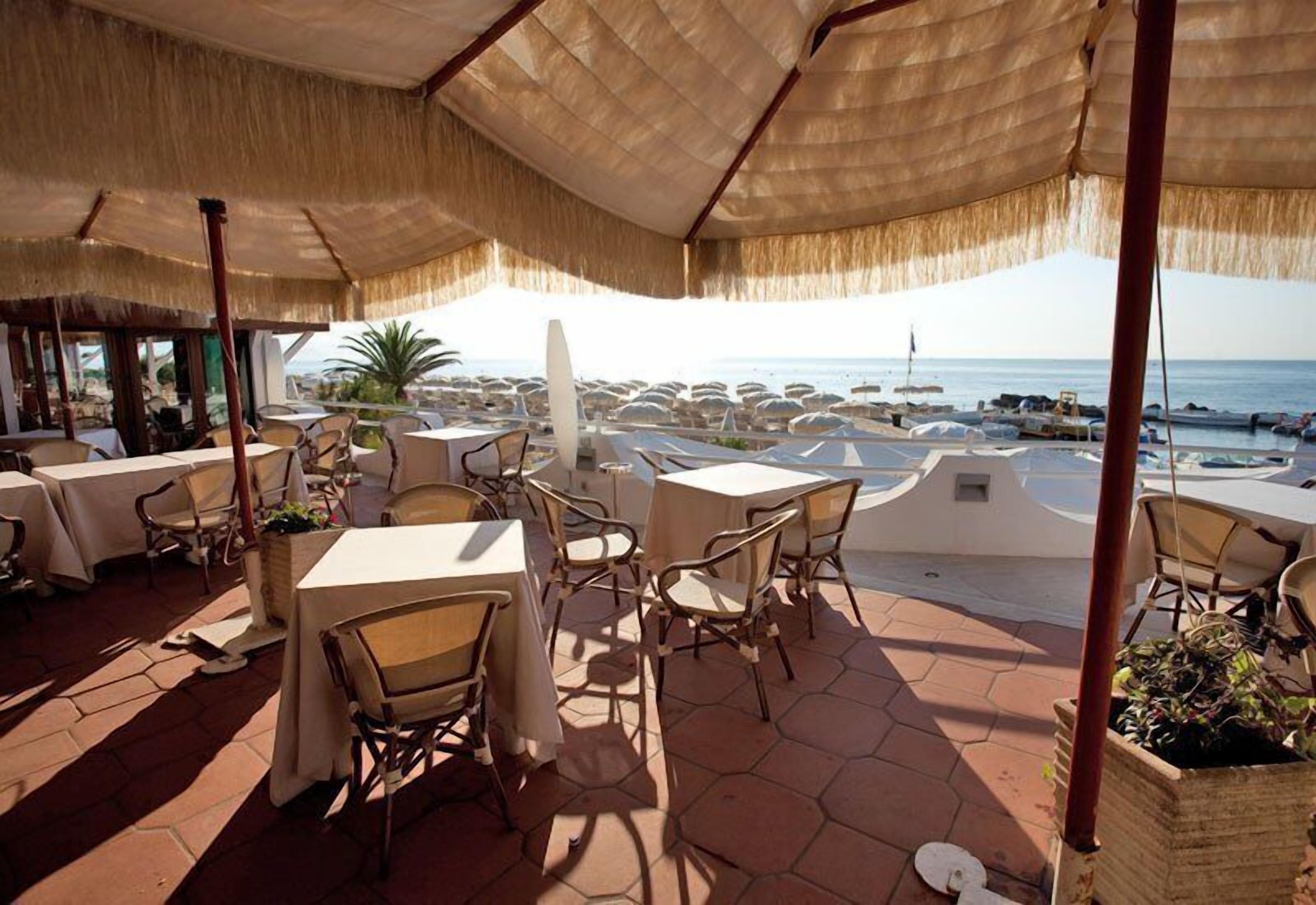 An outdoor dining area with tables and chairs, a thatched roof, and a view of the sea.