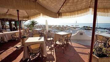 An outdoor dining area with tables and chairs, a thatched roof, and a view of the sea.