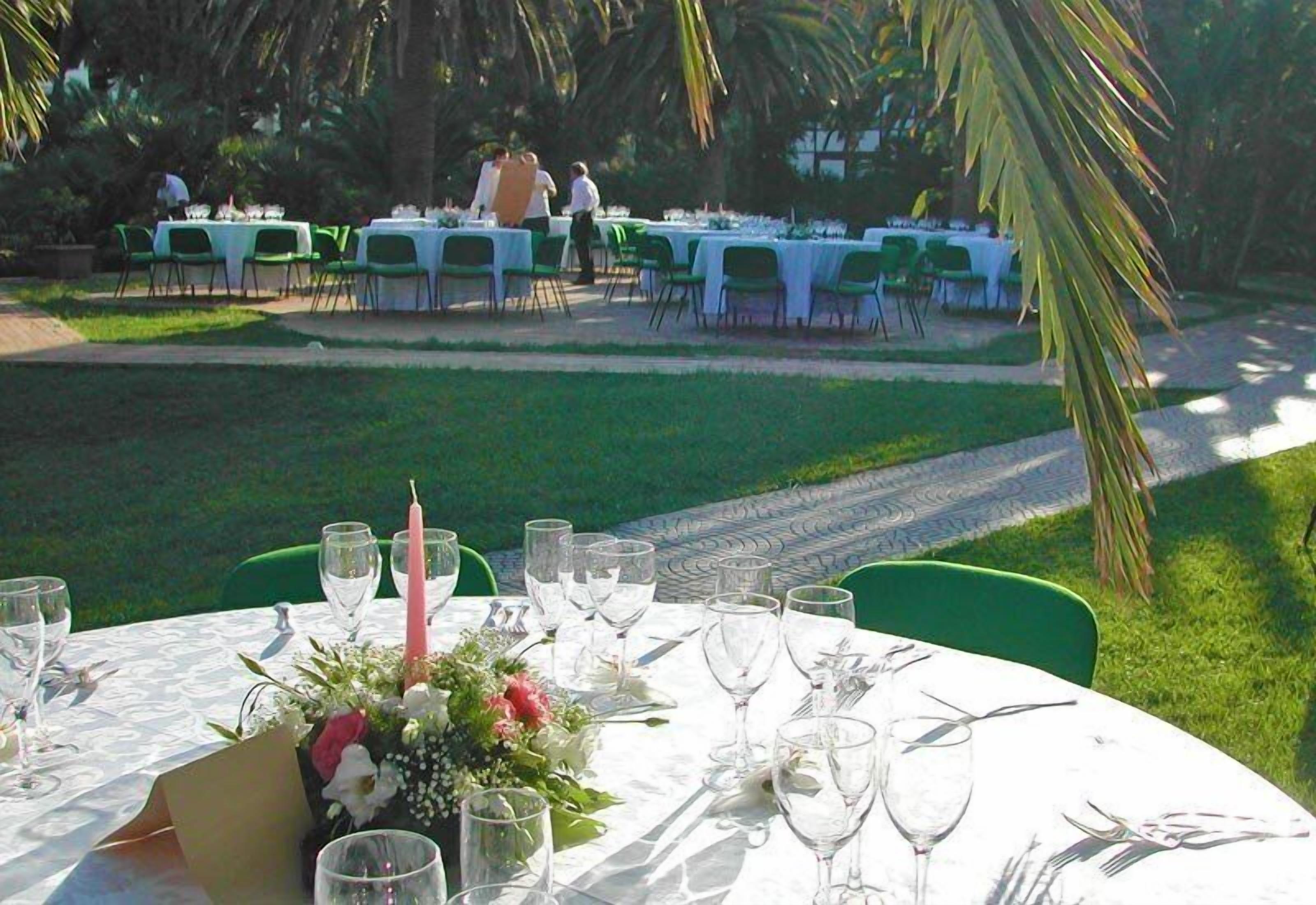 A dining area with round tables set for guests, surrounded by green chairs and palm trees.