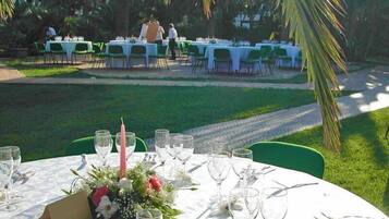 A dining area with round tables set for guests, surrounded by green chairs and palm trees.