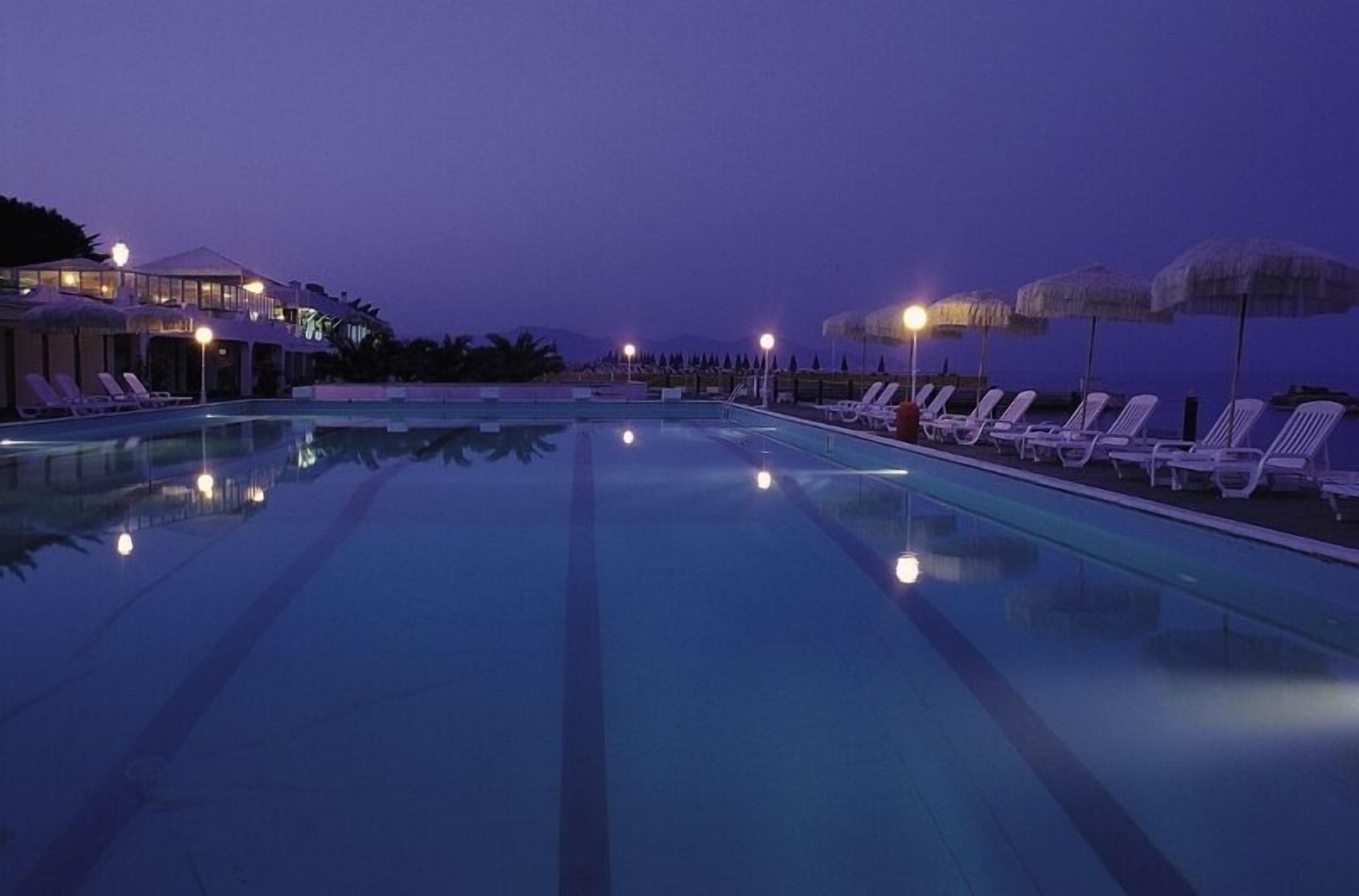 Una piscina con sillas de descanso y sombrillas, rodeada de edificios con luces encendidas.