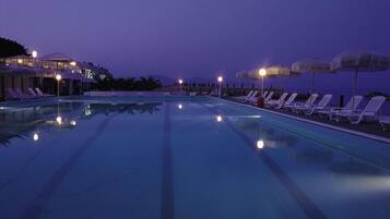 Una piscina con sillas de descanso y sombrillas, rodeada de edificios con luces encendidas.
