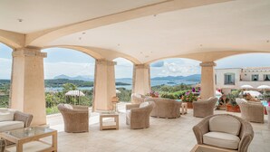 Un espacio de estar techado al aire libre con mobiliario de mimbre, una mesa de centro de vidrio y vistas al mar y las montañas.