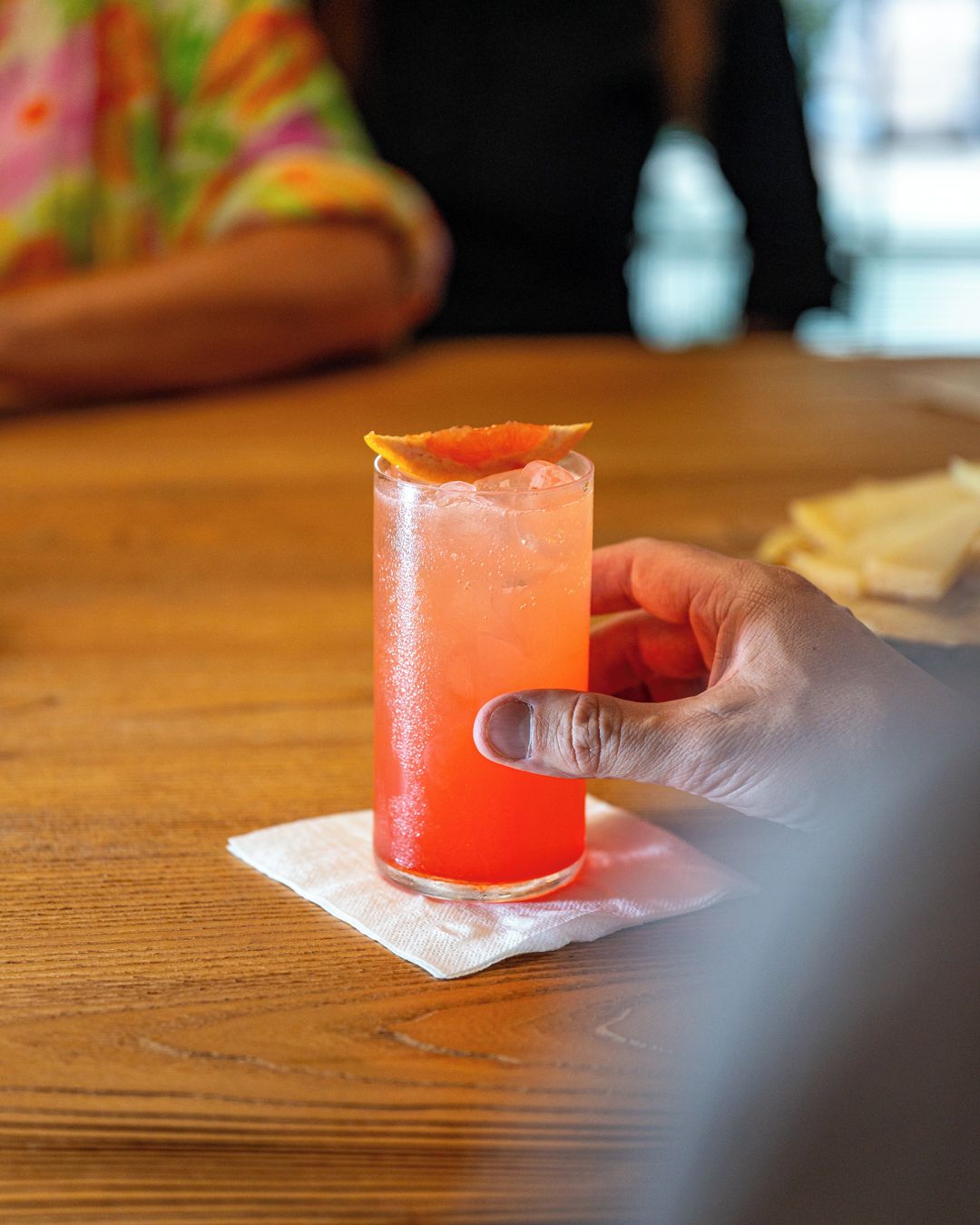 A hand holding a tall glass with a red beverage, ice cubes, and an orange garnish on a wooden table.
