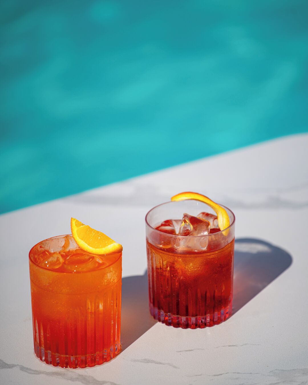 Two glasses of iced tea with orange slices on a white surface near a pool.