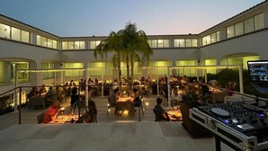 An outdoor dining area with multiple tables and chairs, surrounded by a white building with large windows. - Hotel San Teodoro (San Teodoro)