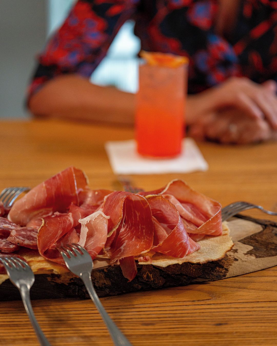 A table with a plate of cured meat and a glass of red beverage.