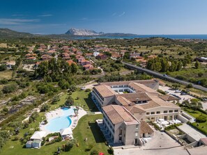 An aerial view of a resort with a swimming pool, a large building with multiple floors, and a mountainous backdrop. - Hotel San Teodoro (San Teodoro)