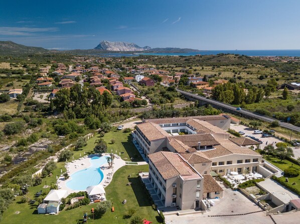 Vista aérea de un complejo turístico con piscina, un edificio grande de varios pisos y montañas al fondo.