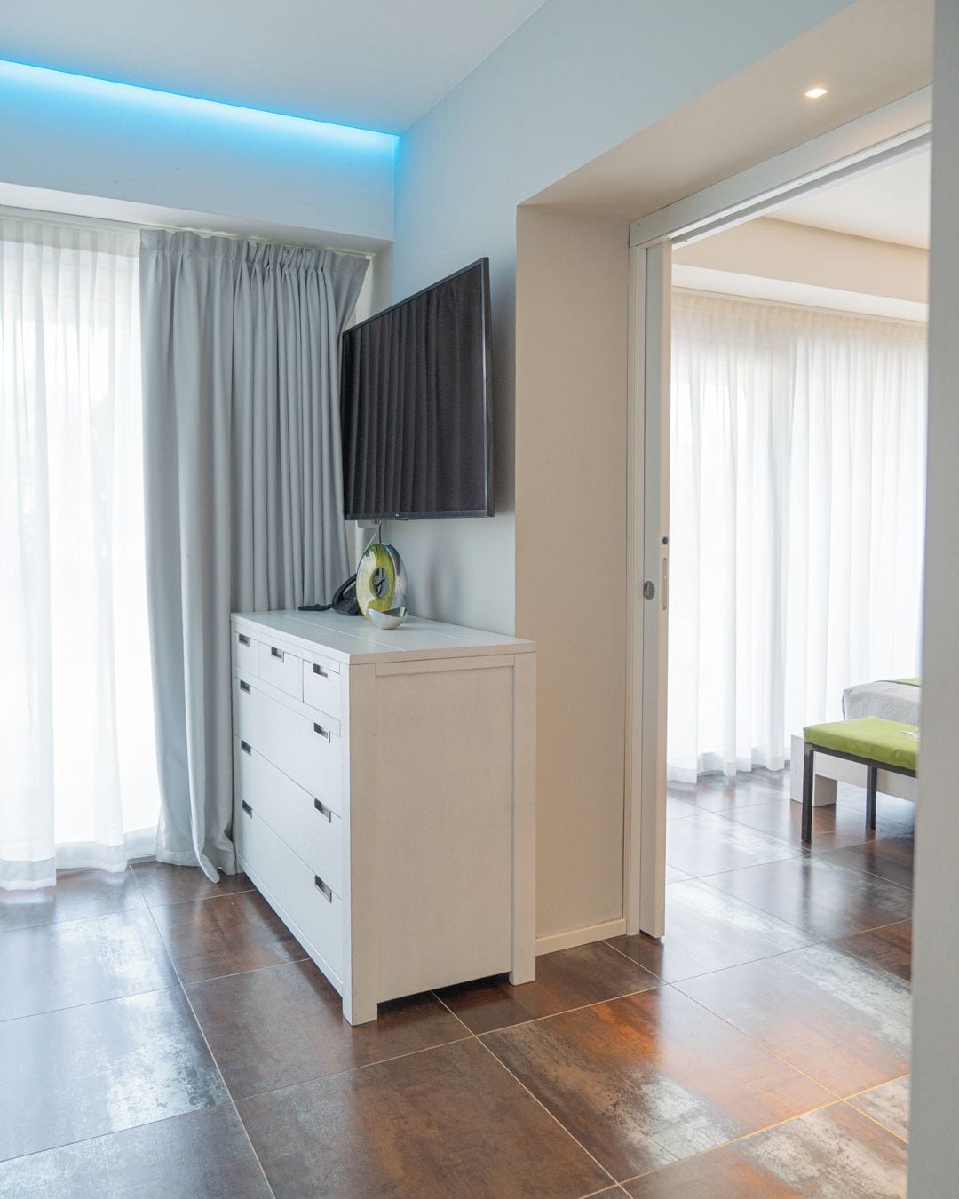 A modern hotel room with a flat-screen TV mounted on the wall, a white dresser, and a large window with curtains.
