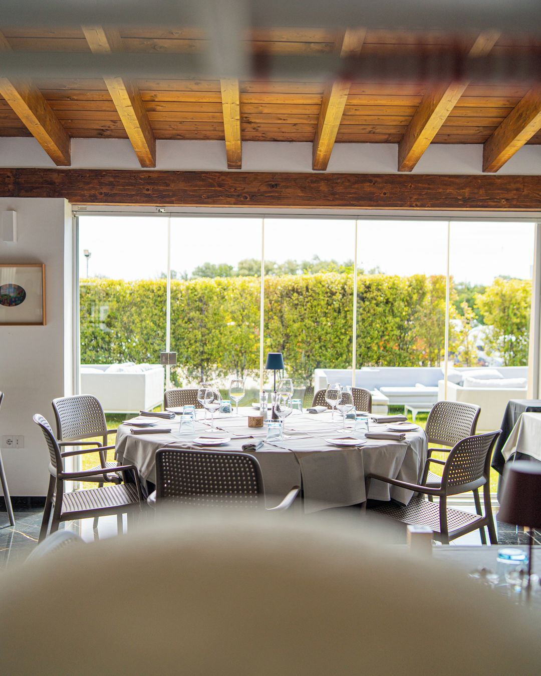 A dining area with a table set for four, surrounded by chairs, and large windows offering a view of a hedge and trees.