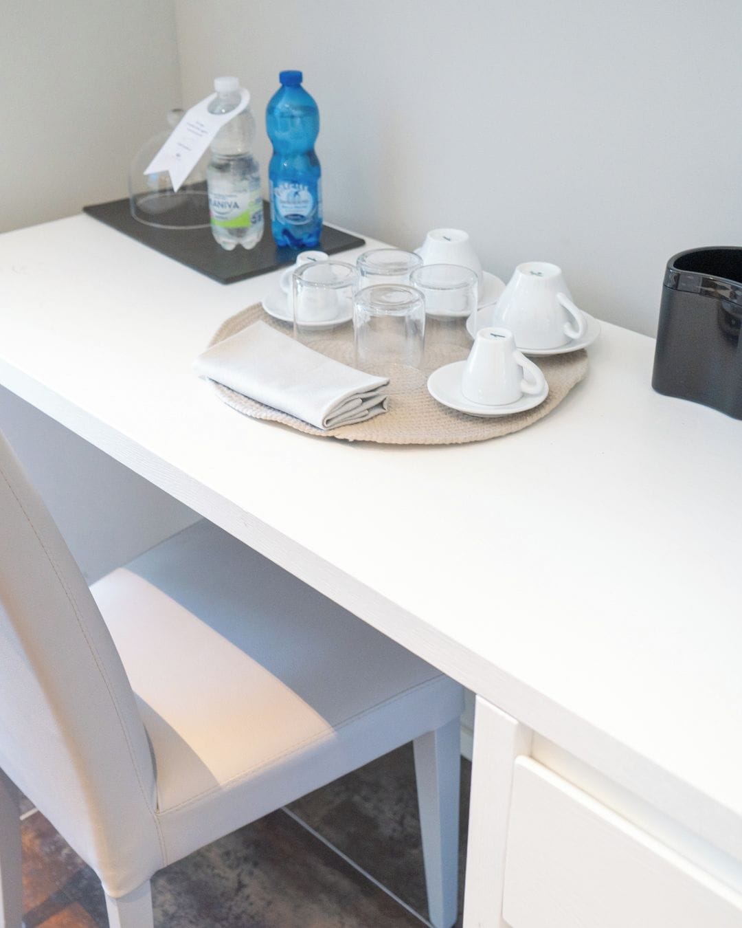 A white desk with a tray holding a folded white towel, a water bottle, a glass, and a cup.