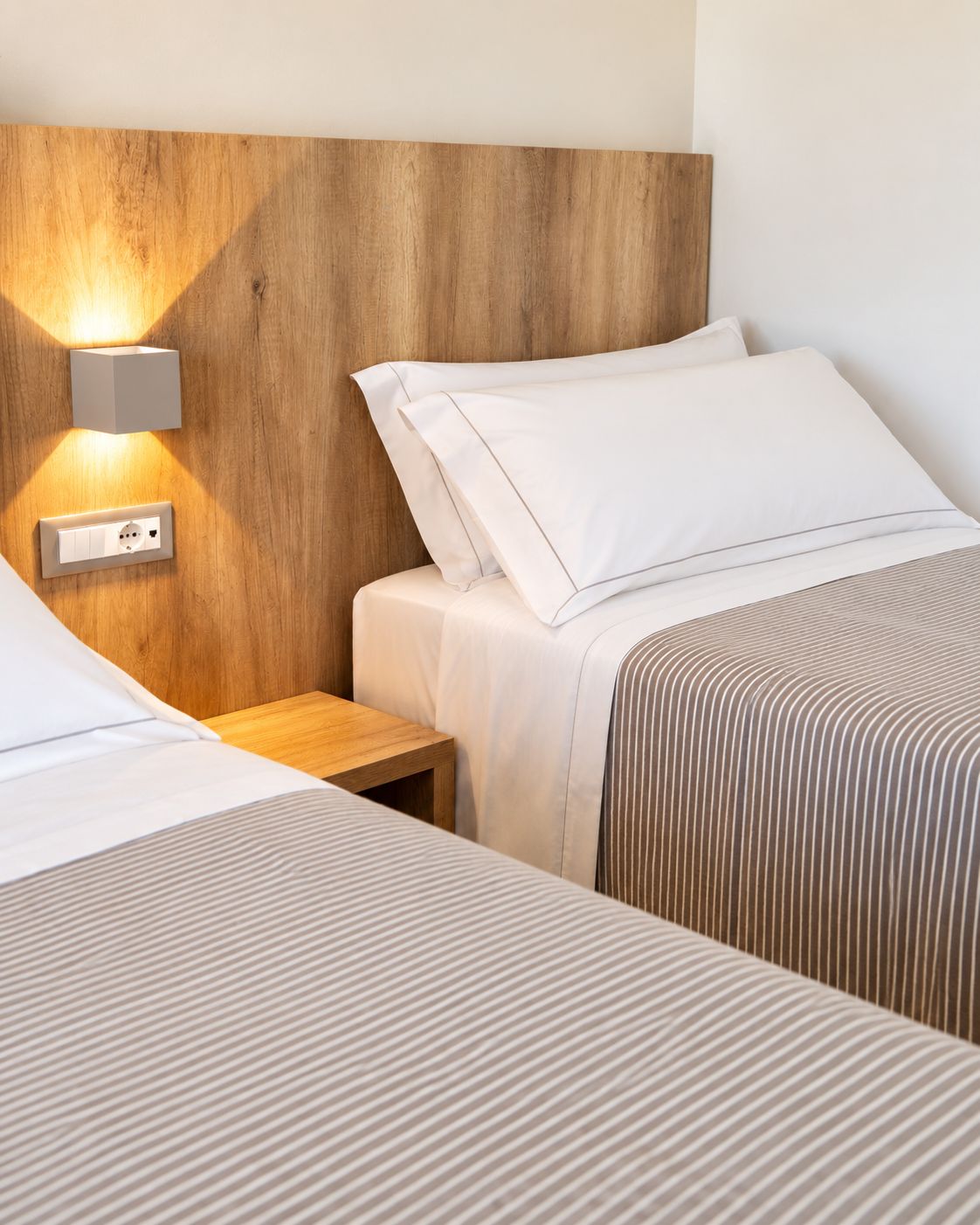 A hotel room with two beds, a wooden headboard, a bedside table, and a wall-mounted light fixture.
