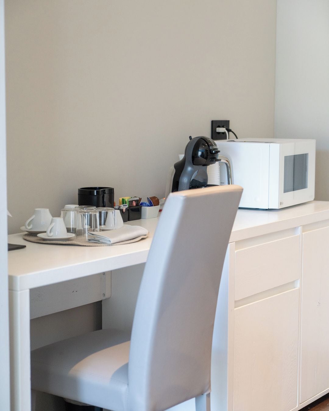 Ein weißer Schreibtisch mit einer Mikrowelle, einer Kaffeemaschine sowie verschiedenen Tassen und Bechern.