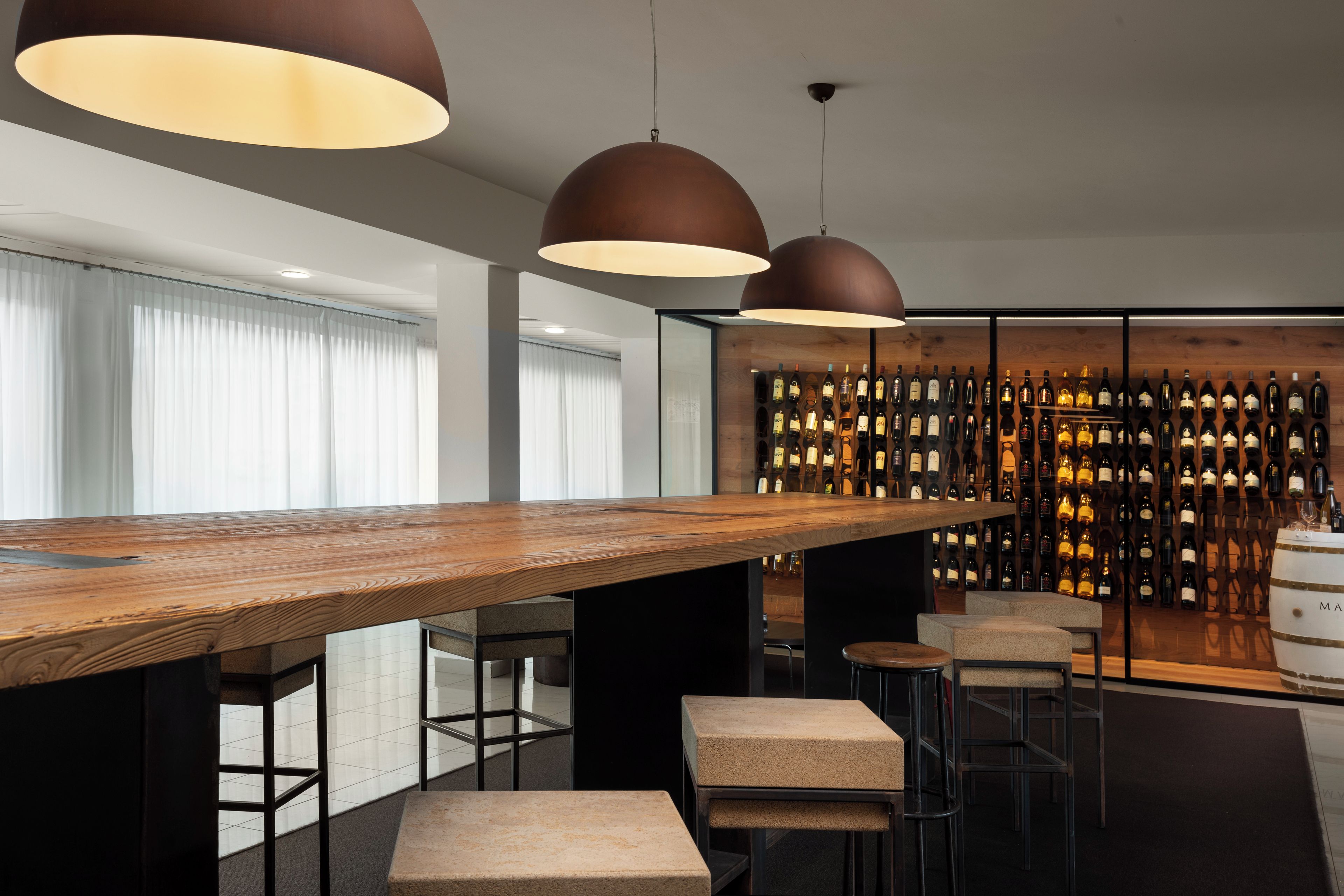 Een moderne bar met een lange houten tafel, meerdere krukken en een wand vol wijnflessen.