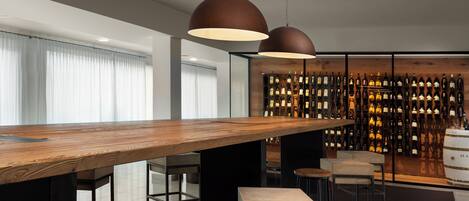 A modern bar area with a long wooden table, multiple stools, and a wall of wine bottles.