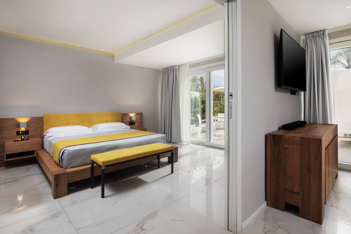 Ein modernes Schlafzimmer mit Bett, Nachttischen, einer Bank, einem Fernseher und einer Schiebeglastür zum Balkon.