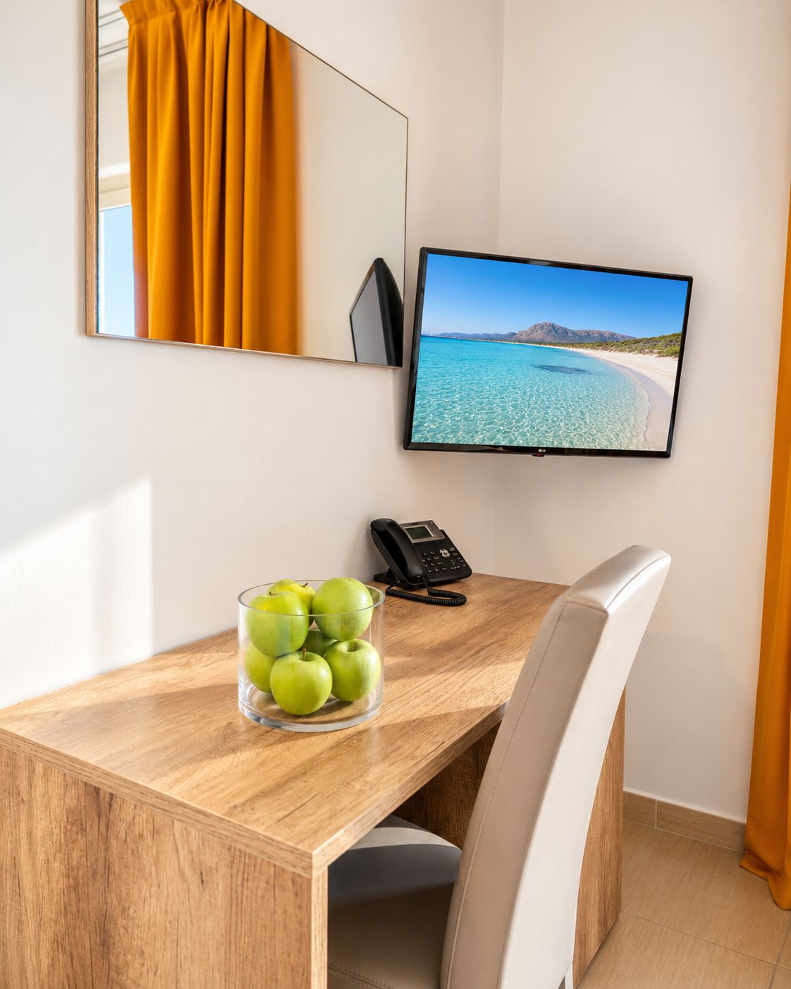 Hotellihuone, jossa on puinen työpöytä, lasivaasi vihreitä omenoita, musta puhelin ja litteänäyttöinen televisio, jolta näkyy kuva rannasta.