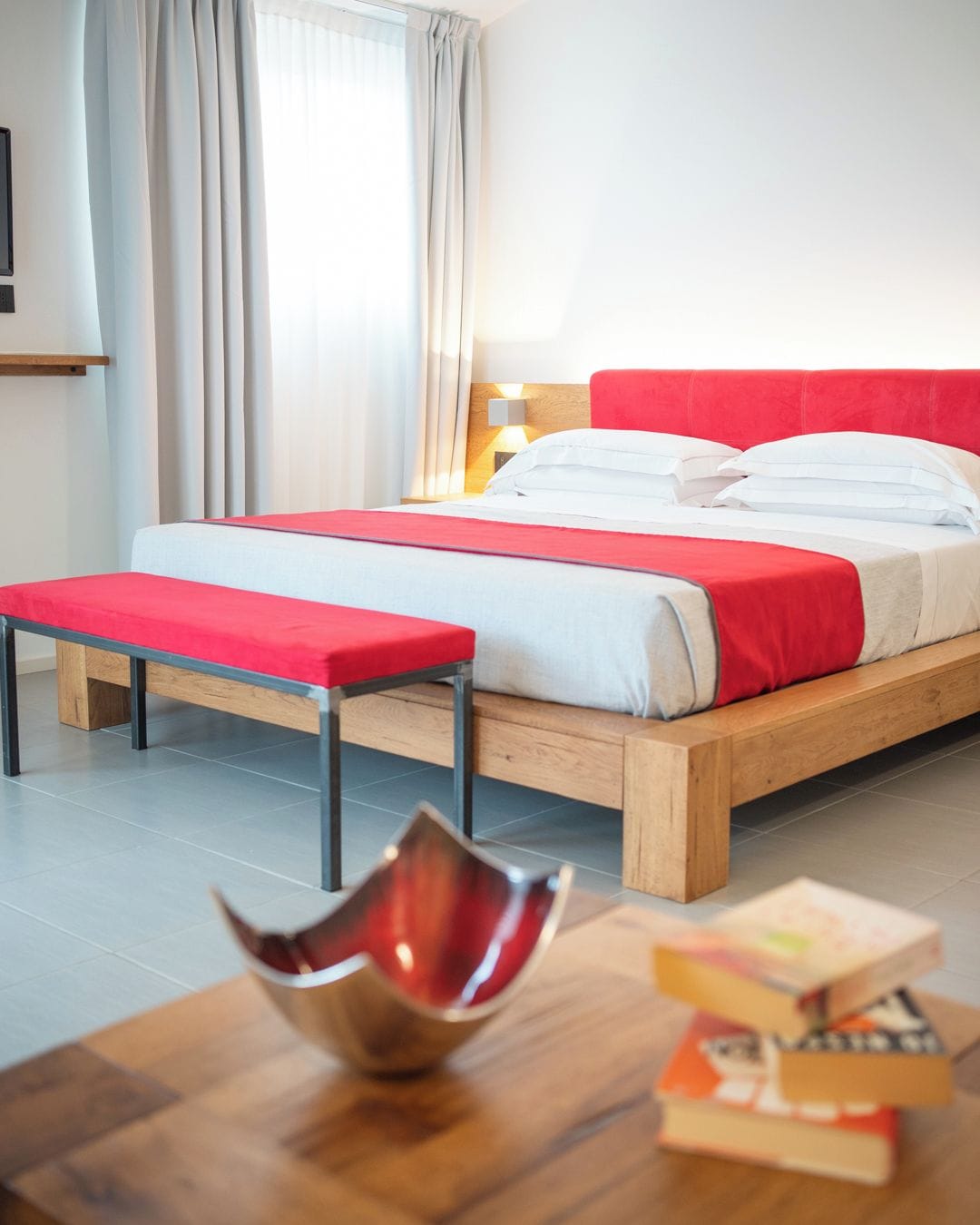 Ein modernes Hotelzimmer mit einem hölzernen Bett, roten Akzenten, einem Nachttisch mit Büchern und einem roten Sessel.