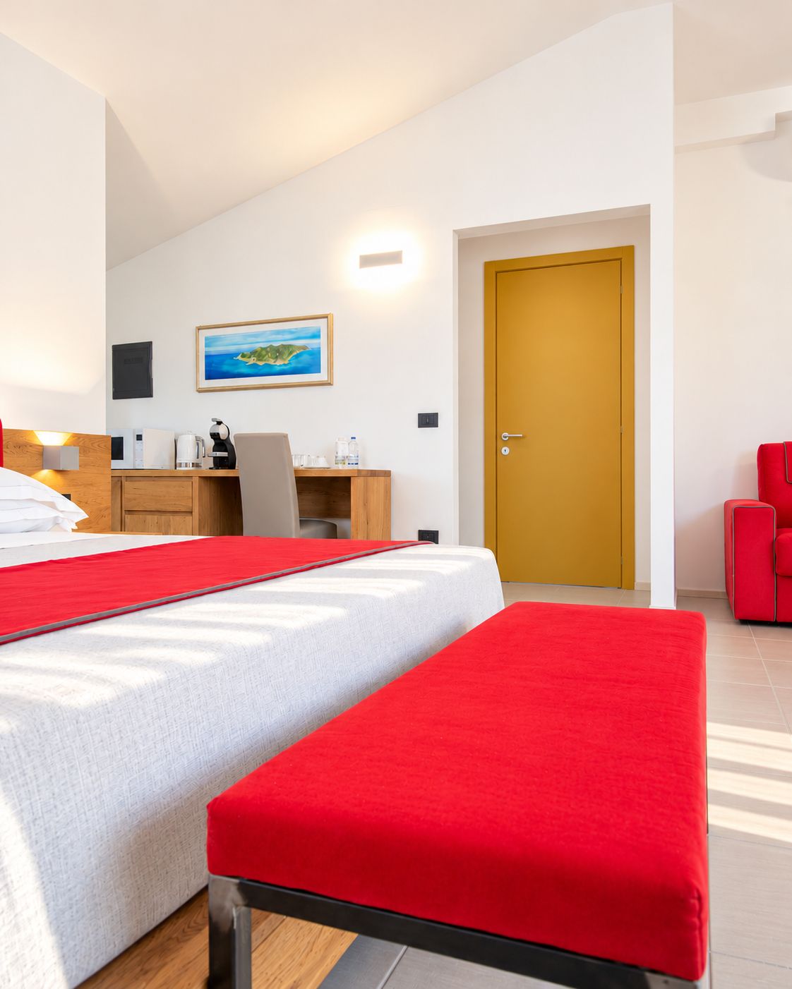 A hotel room with a bed, a desk, a chair, a red bench, and a door.