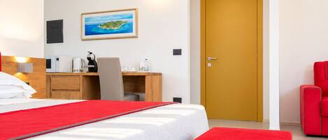 A hotel room with a bed, a desk, a chair, a red bench, and a door.