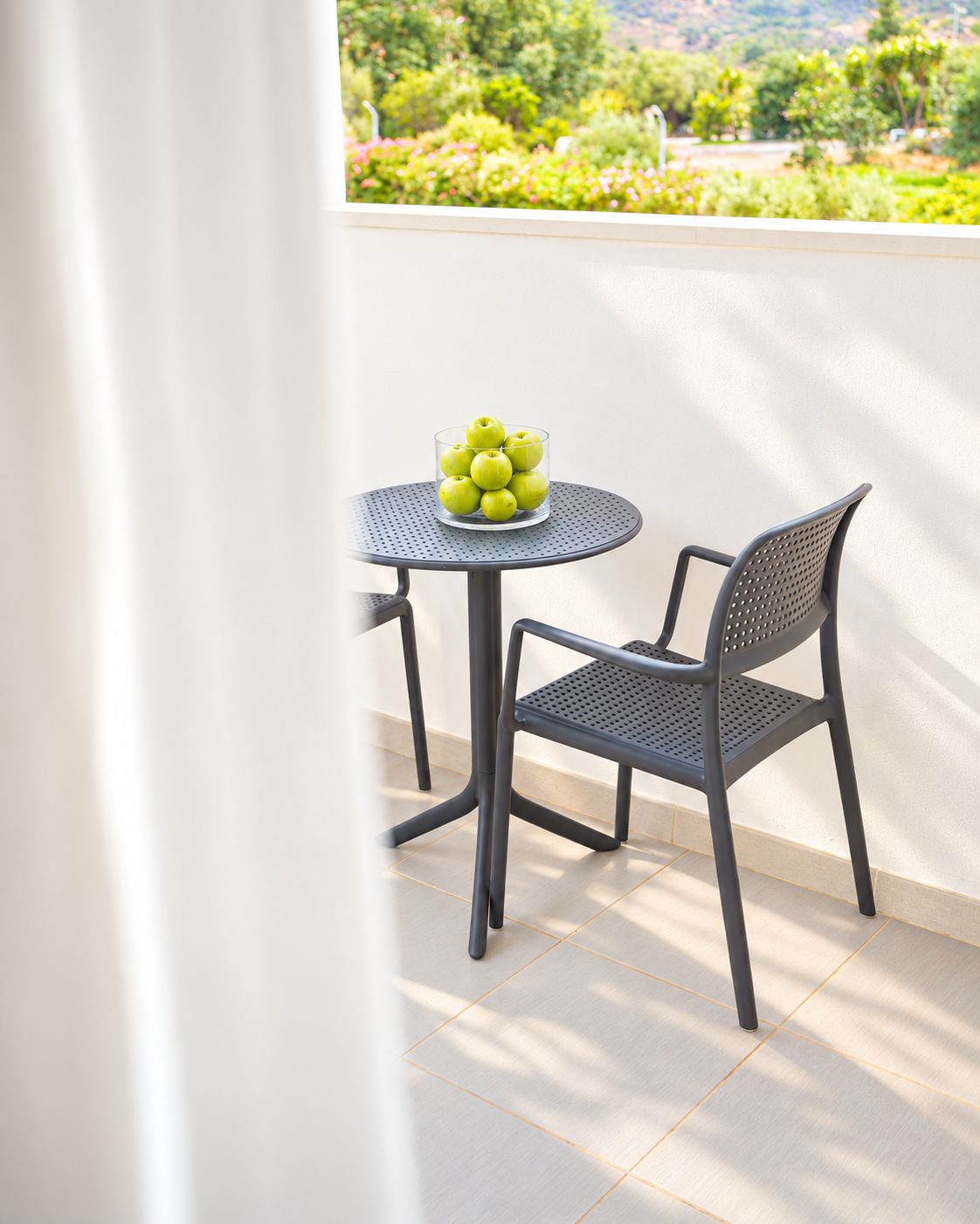 A small round table with a glass bowl of green apples, a metal chair, and a view of a garden with trees and flowers.