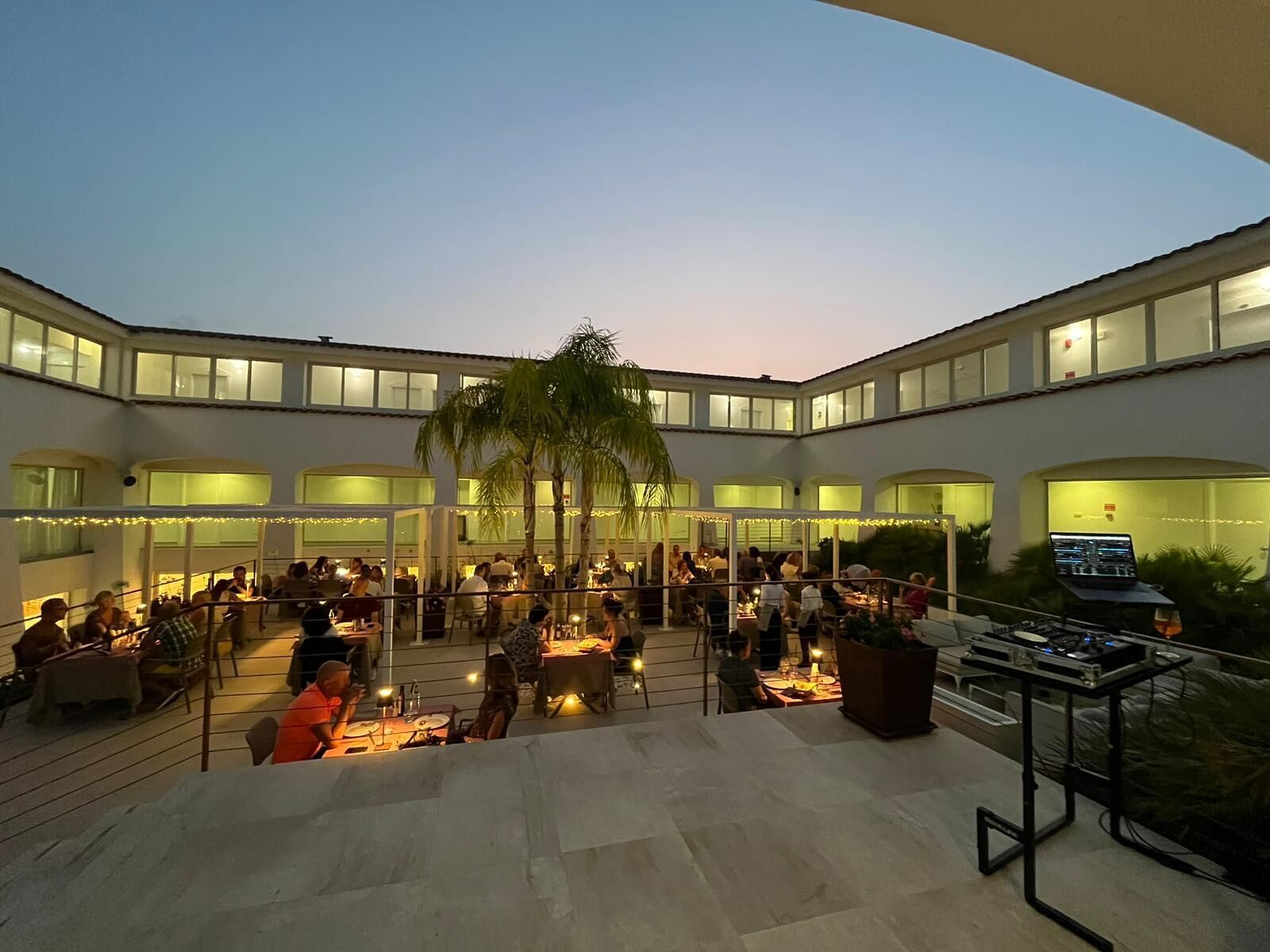A spacious outdoor dining area with multiple tables and chairs, surrounded by a modern building with large windows and a palm tree.
