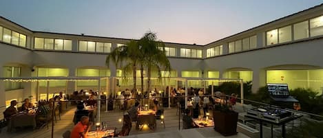 A spacious outdoor dining area with multiple tables and chairs, surrounded by a modern building with large windows and a palm tree.