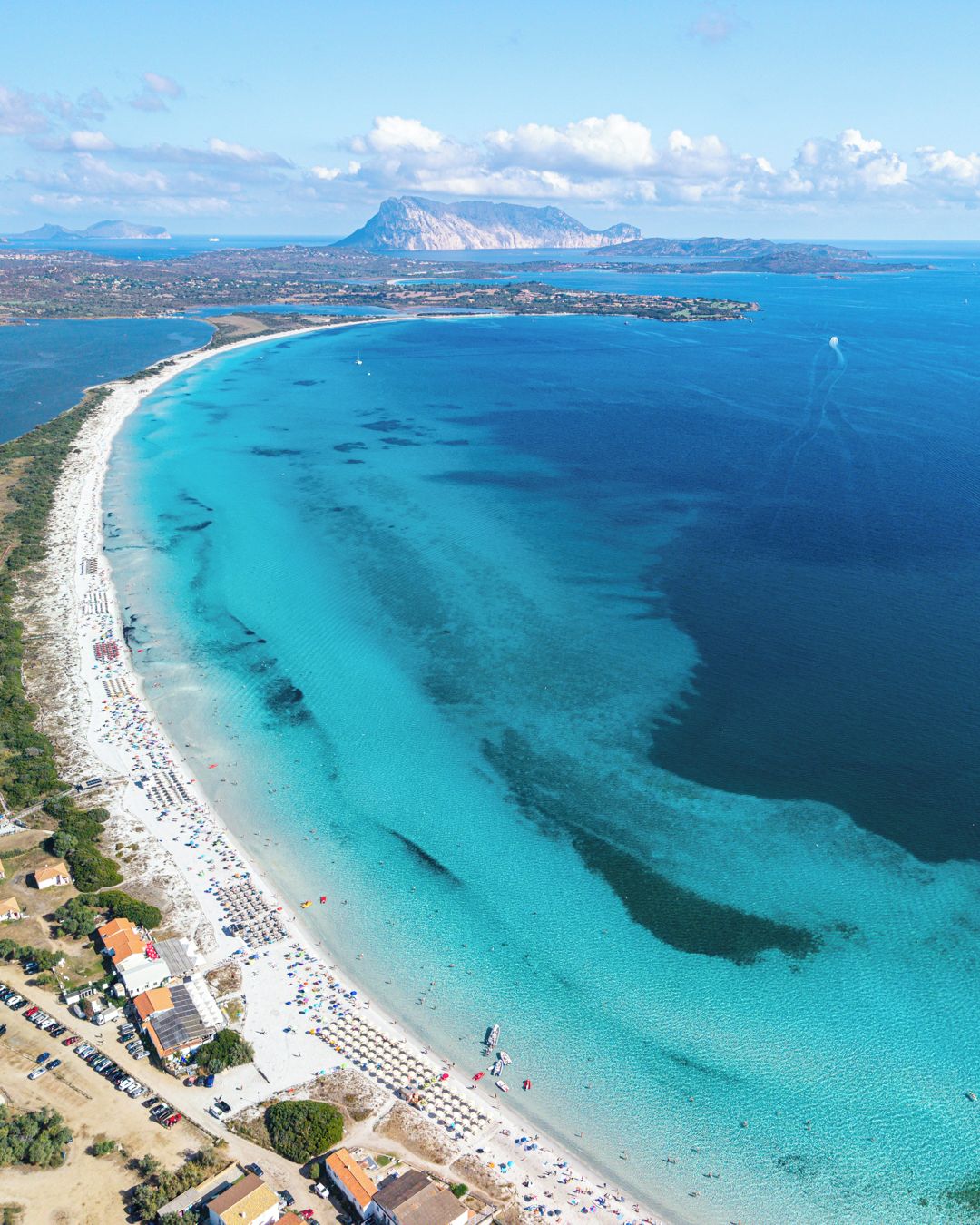 Vista aérea de una zona costera con playa, aguas cristalinas y una montaña al fondo.