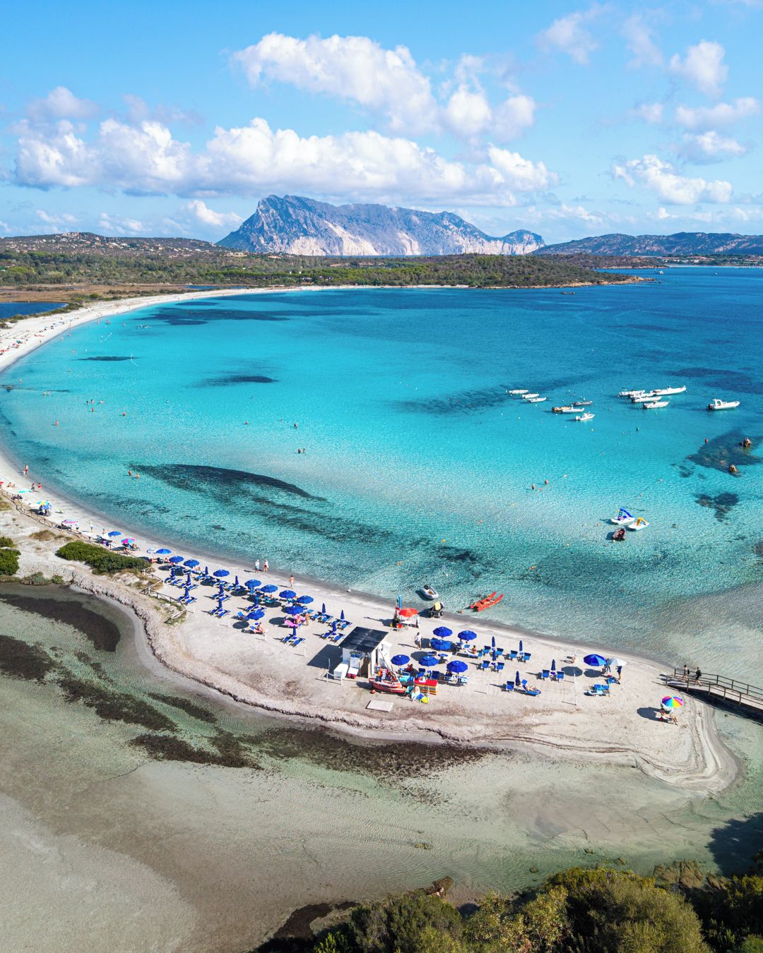 A coastal beach with clear turquoise waters, a sandy shore, and a mountain range in the background.