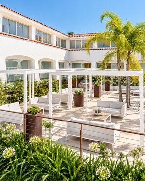 A covered outdoor seating area with white furniture and planters, surrounded by greenery and palm trees. - Hotel San Teodoro (San Teodoro)
