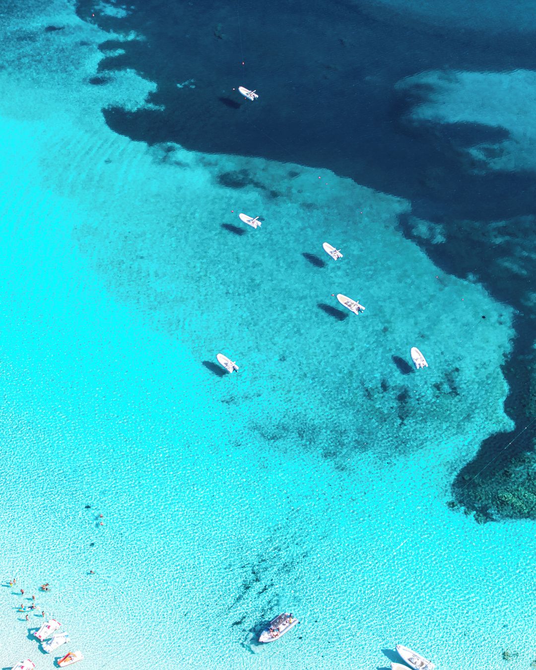 Vista aérea de una playa con aguas turquesas cristalinas, varios barcos y algunas personas.