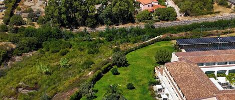 Vista aérea de una zona residencial con piscina rodeada de sillones y sombrillas, un edificio grande con tejado de tejas rojas y montañas al fondo.